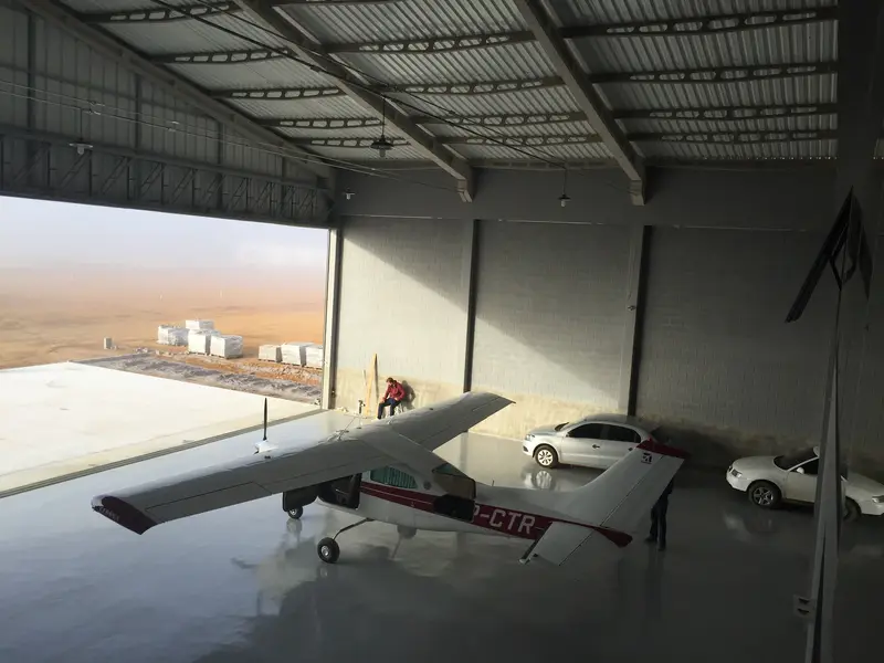 Interior do hangar da Via Rio com um avião monomotor e carros estacionados, portão aberto para a pista