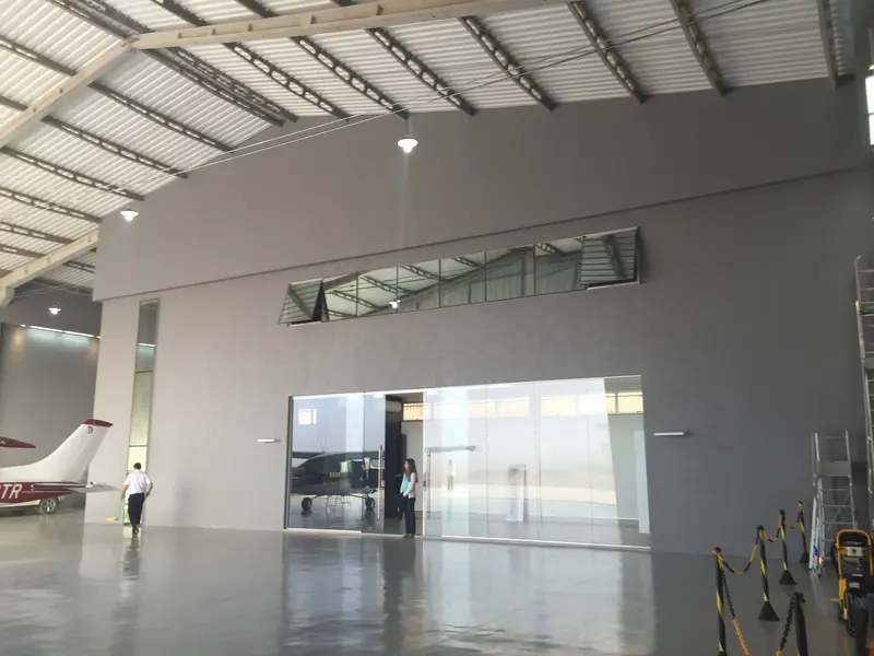 Área interna do hangar, com piso claro, escritório envidraçado ao fundo e a cauda de um avião à esquerda